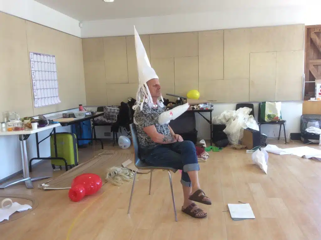 Photograph from Kneehigh's Ubu. Features Mike Shepherd sat on chair in middle of a room wearing tall pointed, white hat. Various props scattered around room in a rehearsal setting.