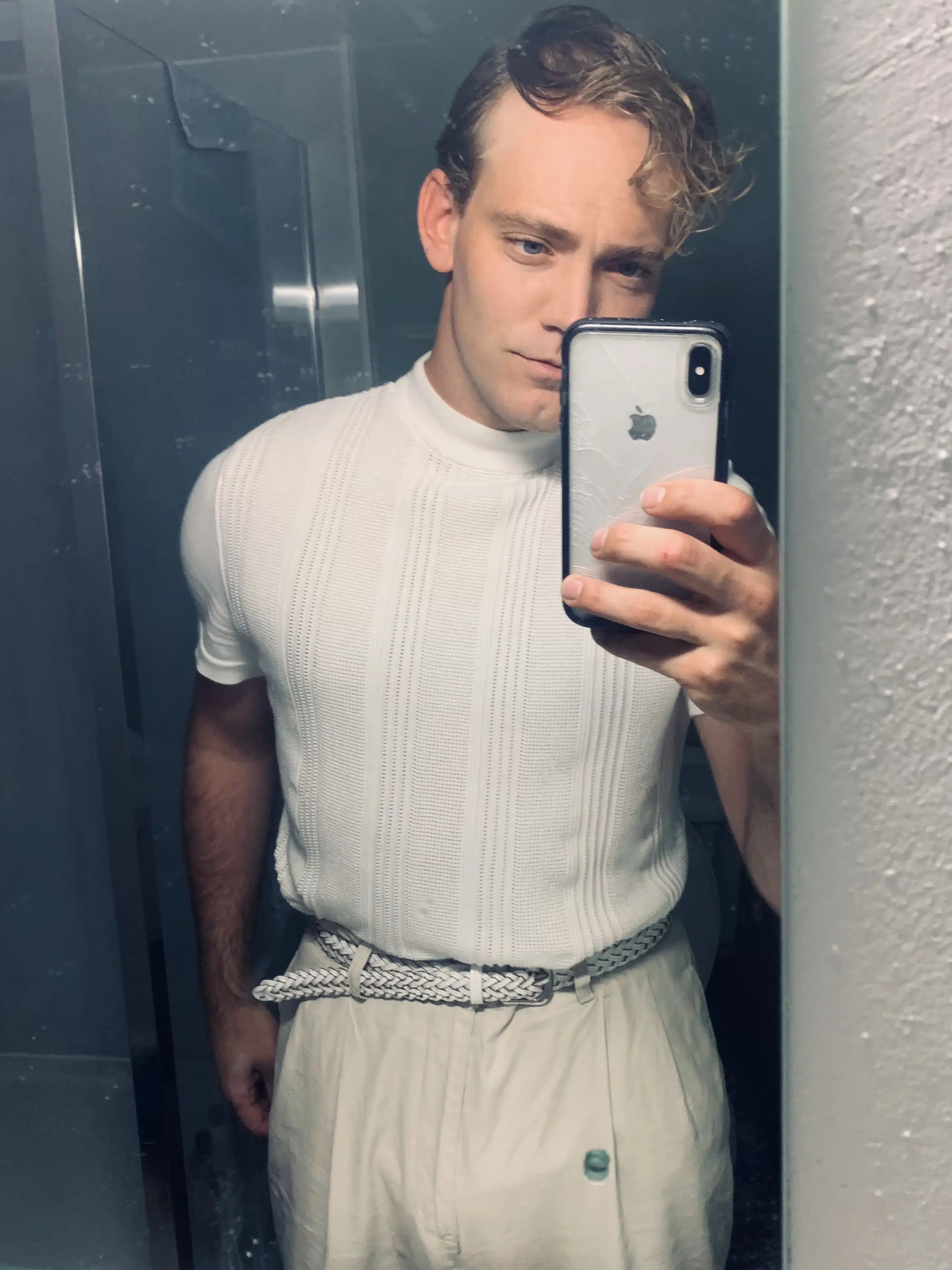 Selfie of Tom Jackson Greaves. The image features an individual standing close in front of a mirror with a white Iphone in their hand. They are dressed in plain white clothing and have long combed over hair.