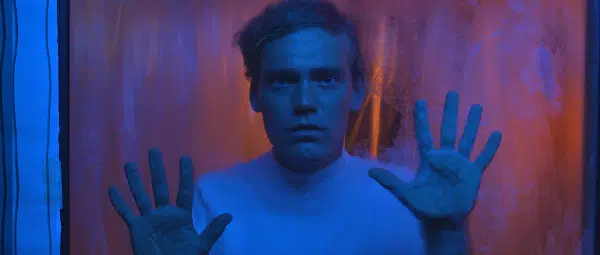 Image of Tom Jackson Greaves. The image features a shot of an individual from the chest up with their hands pressed against a piece of glass in front. They look serious and are illuminated by a blue light.