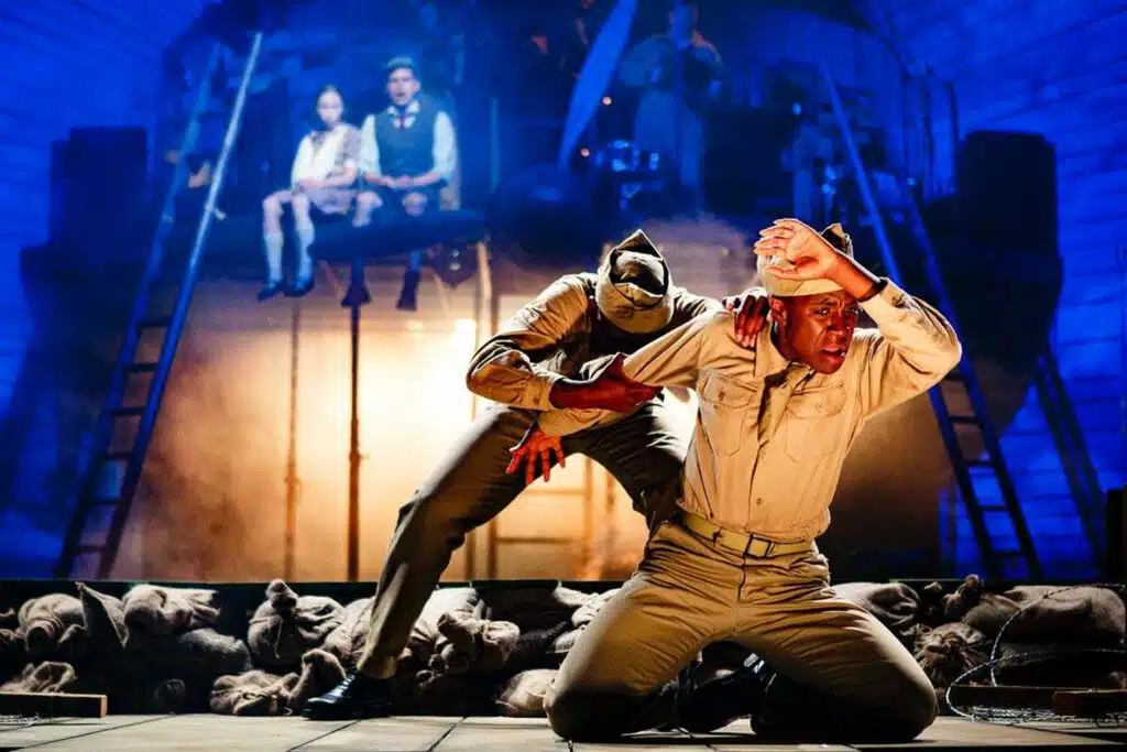 Production photograph. Showing two actors on stage dressed in light Khaki US soldiers uniforms. To the rear, a raised stage with an aircraft propeller at the centre.
