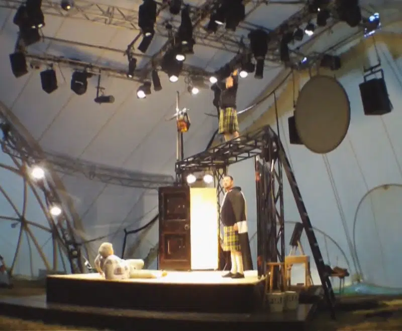 Featured image for Blast at the asylum trailer. The image is blurry and features a small stage inside of a large white dome. There are dozens of stage lights positioned on the ceiling, pointing in different directions. There are three male actors on the stage in the centre of the image. Two are dressed in tartan kilts, with one standing on a metal structure high above the stage. They are both looking at the third actor who is dressed in white and laying on the stage floor. A small section of the audience can be seen in the bottom left of the image.