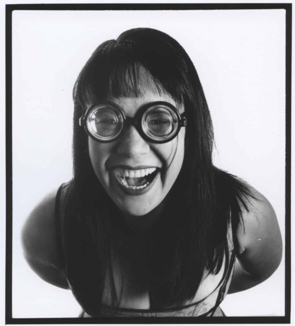 Black and white photograph of a woman, leaning into the camera. Cropped to show her head and torso. She has long straight dark hair with a fringe, and wears a low cut top, although her chest is most obscured by her hair. She is smiling, and wears goggle style oversized glasses.