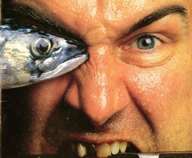 Programme features a close set photograph of a man's face. His right eye is obscured by the head of a mackerel and he holds a knife between his teeth - one of which is gold. His composure appears angry.
