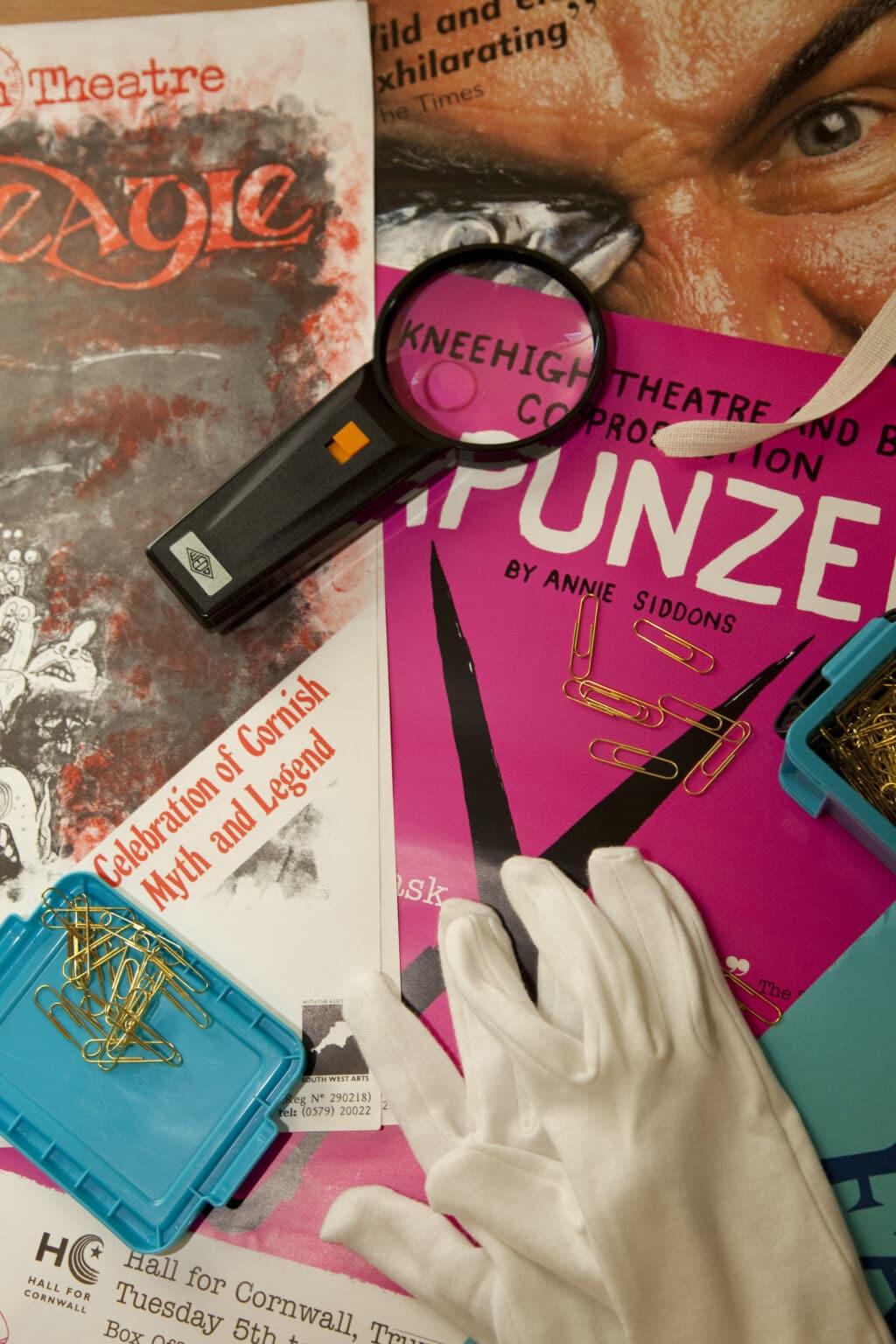 A selection of Kneehigh posters, overlaid with white cotton gloves, a magnifying glass and box of brass paperclips