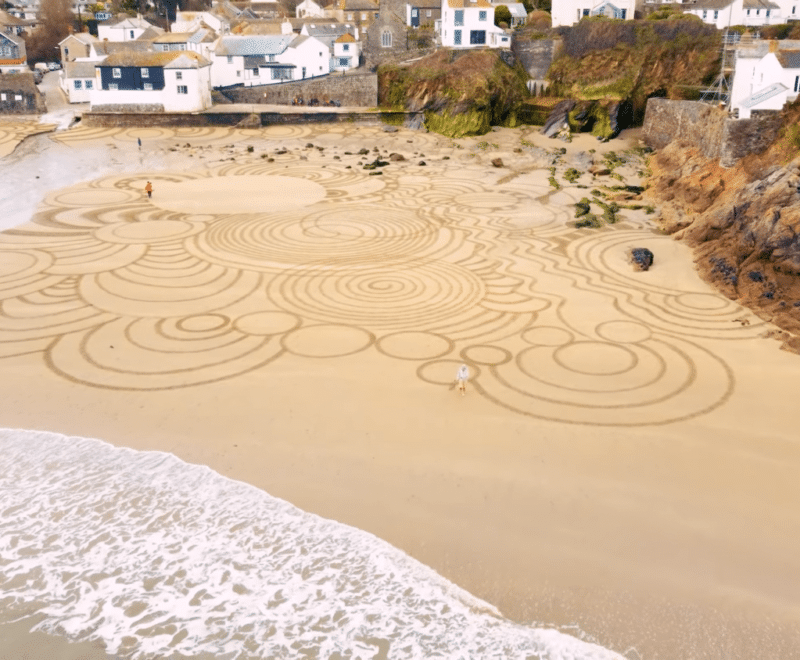 Thumbnail image from Random Acts of Art video. An aerial shot of a beach in St Austell bay taken in the daytime with elaborate circular marks made in the sand. Houses and cliffs are also visible in the periphery.