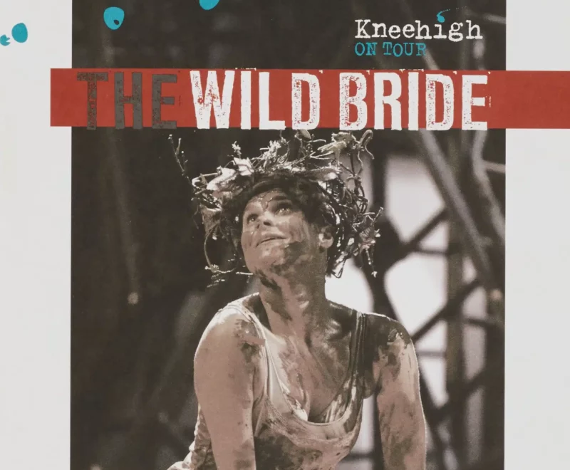Features a production image of a woman wearing a head-dress made of leaves and twigs covered in mud and kneeling on stage. The production title 'The Wild Bride' is presented above the image in black and white font set against a red banner.