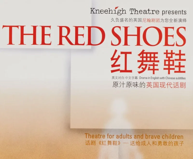 Image features a pair of digitally edited red leather shoes positioned vertically on a wooden floor. The production title 'The Red Shoes' is presented in the top right corner of the page in a bright red font. There are symbols in mandarin presented below the title. Small black text beneath the title reads 'Drama in English with Chinese Subtitles'. A separate piece of text half way down the page reads 'Theatre for adults and brave children'.