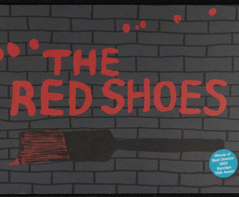 Image features a cartoon illustration of a grey brick wall on which 'The Red Shoes' is written in a red font resembling paint. A cartoon image of a paint brush is positioned below the text. A small blue circle in the bottom left corner contains white text that reads 'Winner of Best Director 2002 Barclays TMA Award'