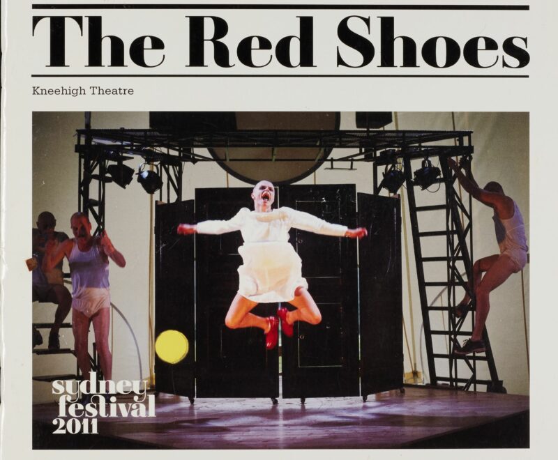 Image features a colourful production photograph in the centre of an off white backdrop. The photograph features a female performer in a white dress and red shoes jumping in a state of excitement on stage. The production title 'Red Shoes' is presented in bold Black text above the image.