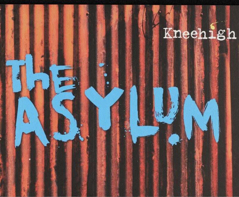 Image features text reading 'The Asylum' in a graffiti style blue text set against the backdrop of a orange rusty corrugated metal fence. The typewriter Kneehigh Theatre logo is positioned in the top right.
