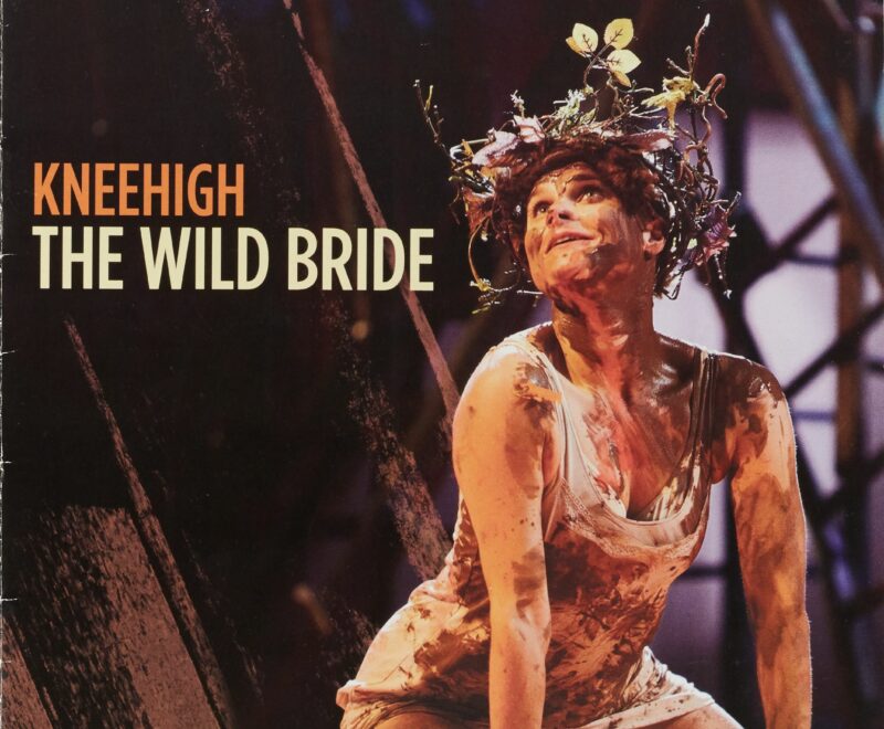 Features a production image of a woman wearing a head-dress made of leaves and twigs covered in mud and kneeling on stage. The production title is presented in a bold white font to the left of the image. Bold text reading 'St. Ann's Warehouse' is presented at the bottom of the image.