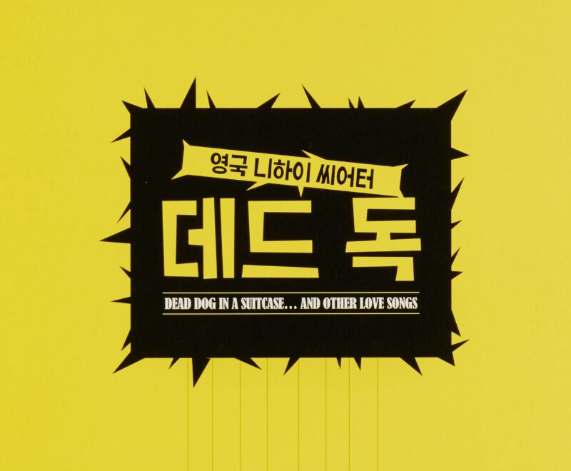 The image features a black square in the centre of a bright yellow backdrop. The square has spikes poking out in all directions and has eight light bulbs suspended by pieces of string. Inside the square is yellow font written in Korean and 'Dead Dog in a Suitcase... and other love songs' in a white font.