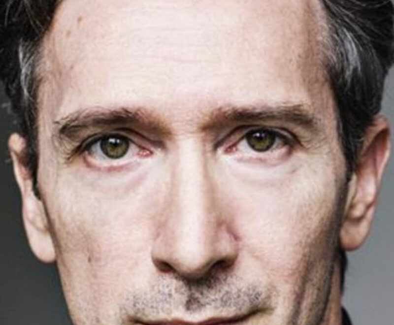 A person with short, gray hair and a serious expression, wearing a black shirt, looks directly at the camera.